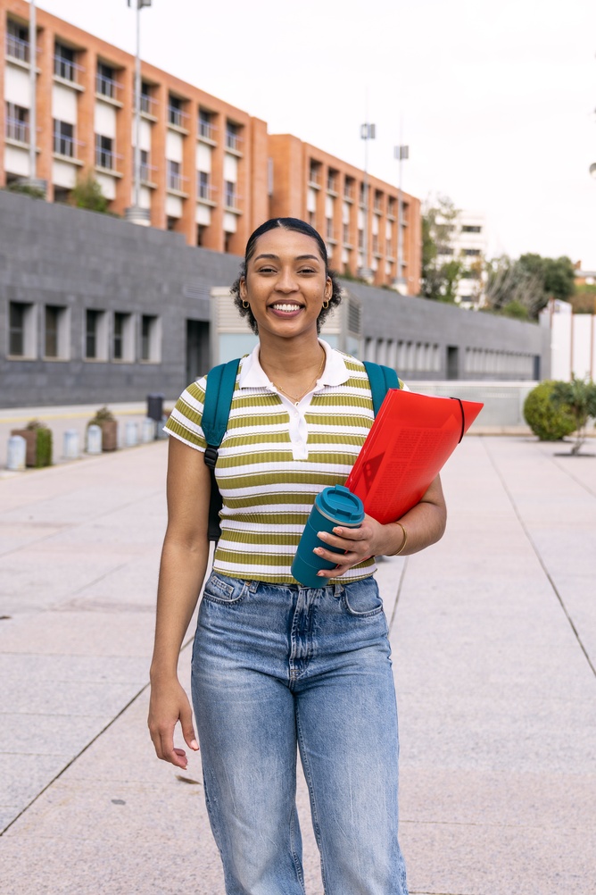 Photograph of a student walking outdoors on a campus pathway with modern buildings in background. The student wears a striped shirt, jeans, and backpack, holding a red folder and blue reusable cup, suggesting a study or school-related context.